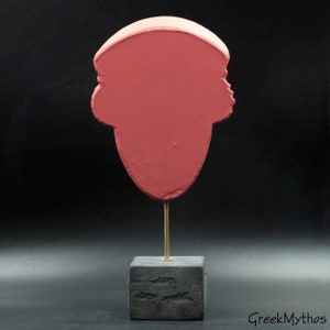 May include: A pink plaster bust of a person's head on a black wooden base with a gold metal stand.