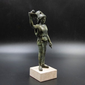May include: A bronze statue of a woman wearing a helmet and holding a bird in her raised hand. The statue is standing on a square base.