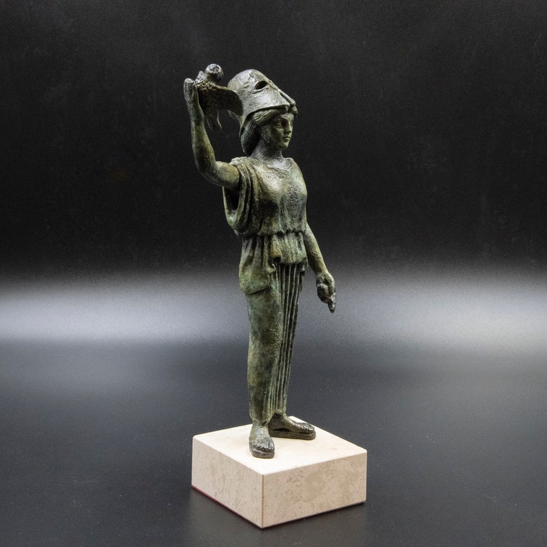 May include: A bronze statue of a woman wearing a helmet and holding a bird in her raised hand. The statue is standing on a square base.