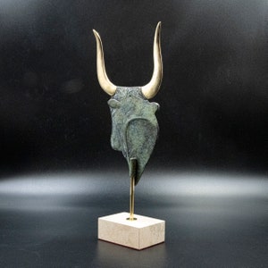 May include: A bronze sculpture of a bull's head with gold horns on a white marble base.
