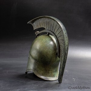 May include: A bronze Greek warrior helmet with a crested plume. The helmet is detailed with a textured surface and a decorative design.