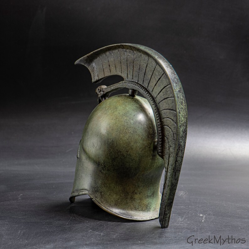 May include: A bronze Greek warrior helmet with a crested plume. The helmet is detailed with a textured surface and a decorative design.