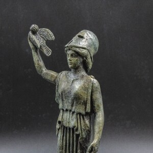 May include: A bronze statue of a woman wearing a helmet and holding a bird in her raised right hand. The statue is detailed and has a weathered finish.