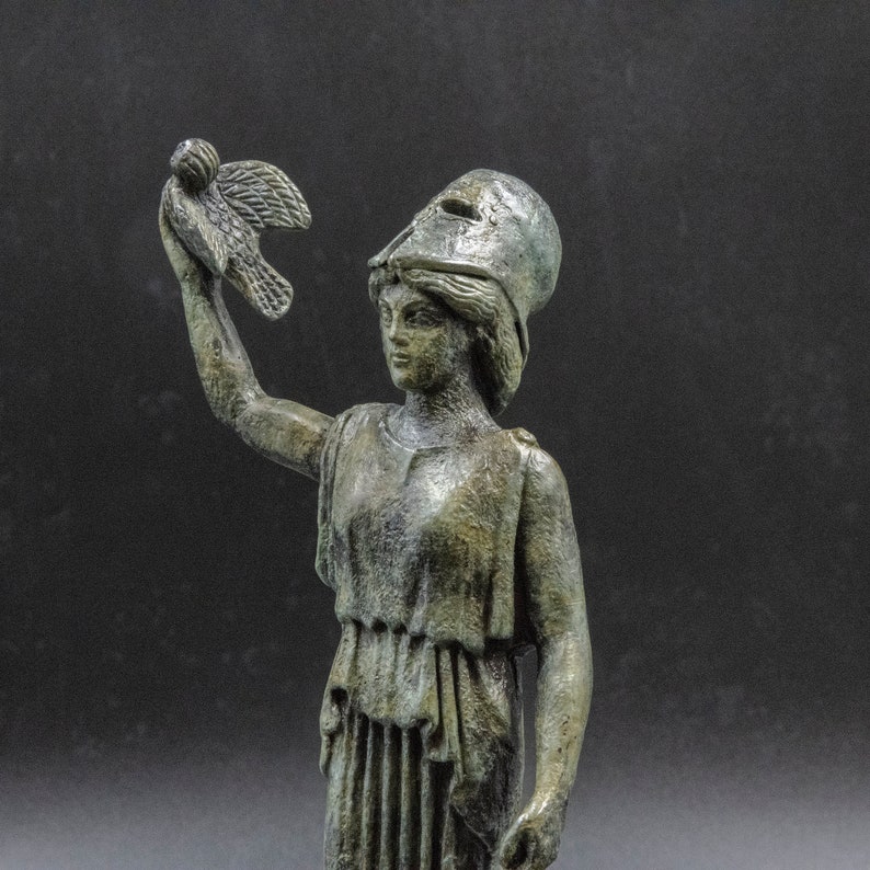 May include: A bronze statue of a woman wearing a helmet and holding a bird in her raised right hand. The statue is detailed and has a weathered finish.