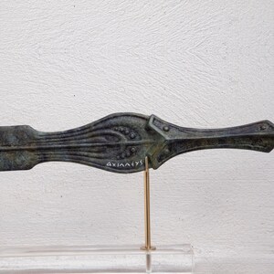 May include: Bronze-colored, antique-style dagger with intricate detailing. The blade features a central ridge with decorative patterns and small circular accents. The handle has a flared design with a rectangular guard. The word "AXIA&Lambda;EYS" is inscribed on the blade.