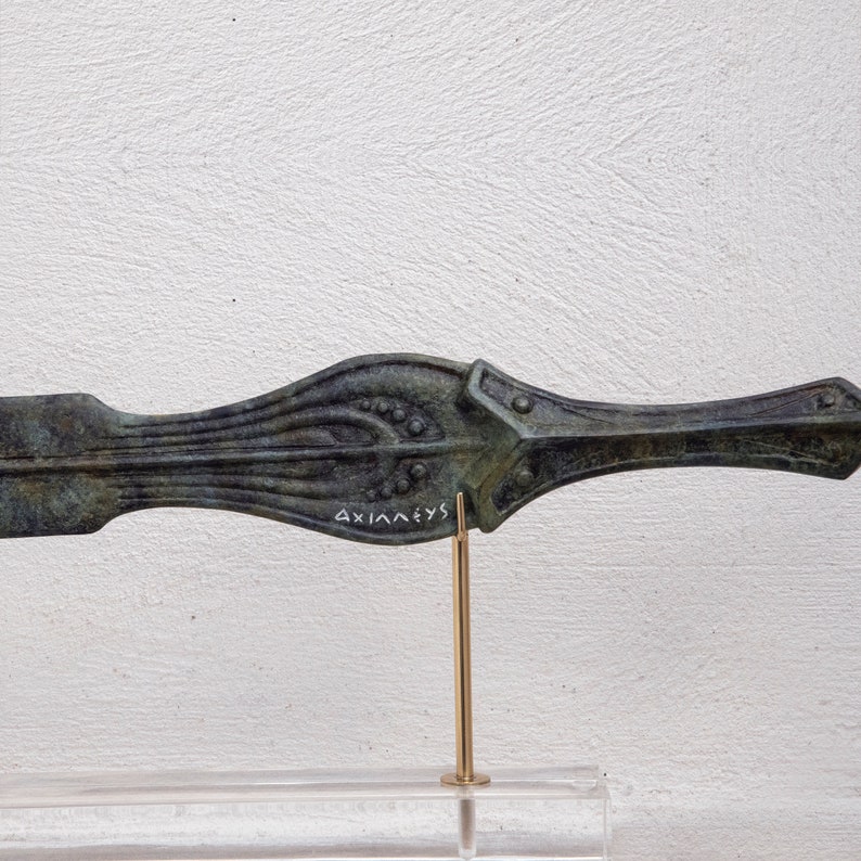 May include: Bronze-colored, antique-style dagger with intricate detailing. The blade features a central ridge with decorative patterns and small circular accents. The handle has a flared design with a rectangular guard. The word "AXIA&Lambda;EYS" is inscribed on the blade.
