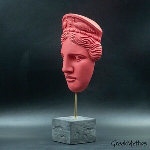 May include: A red painted plaster bust of a woman with a crown on her head. The bust is on a black square base with a gold metal rod.