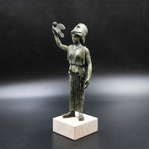 May include: A bronze statue of a woman holding a bird in her outstretched hand. The woman is wearing a helmet and a long, flowing robe. The statue is standing on a square base.