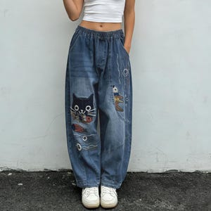 Pode incluir: Calças de ganga azuis com cintura elástica e corte folgado. As calças apresentam aplicações bordadas de um gato preto e peixes. Sapatilhas brancas completam o visual.