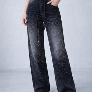 Puede incluir: Vaqueros negros de pernera ancha con cintura con cordón y pedrería dispersa. Los pantalones vaqueros tienen un lavado descolorido y un diseño clásico de cinco bolsillos. Los pedrería se concentran en la parte superior y se desvanecen por las piernas.