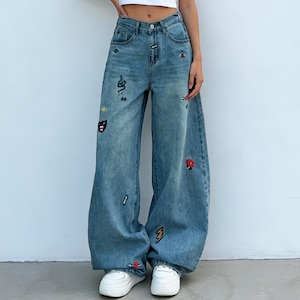 May include: Light blue denim jeans with a wide leg cut. The jeans are decorated with embroidered patches including a snake, lightning bolt, and playing card symbols. They have a classic five-pocket design and a button and zip closure.