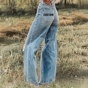 May include: Light blue denim jeans with a high waist and wide legs. The jeans have distressed details, including a ripped knee. The back pocket features the word "CUSTOM" in a black font.