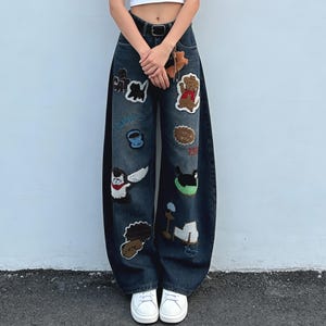 May include: Dark wash denim jeans with a wide leg design. The jeans are adorned with various embroidered patches, including cartoon characters and other whimsical designs. The jeans have a black belt and are paired with white sneakers.