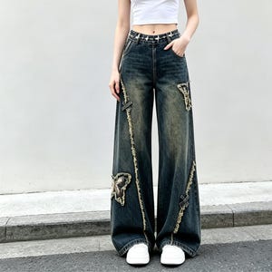Könnte beinhalten: Dunkelblaue Jeans mit weitem Bein und Used-Details. Die Jeans hat dekorative Patches und ausgefranste Kanten. Die Taille ist mit einem Nietengürtel akzentuiert. Die Jeans wird mit weißen Turnschuhen kombiniert.