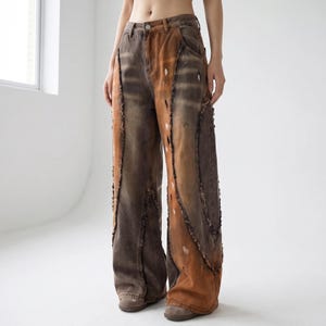 May include: Wide-leg jeans in shades of brown and orange with distressed details. The denim pants feature a high waist and unique paneling with frayed edges. White paint splatters add a touch of artistic flair to the design.