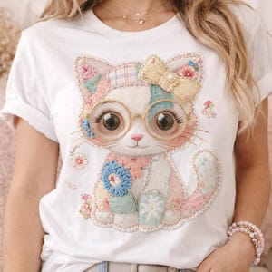 May include: White t-shirt featuring a whimsical cat design. The cat is adorned with glasses, a bow, and patchwork details in pastel colors. Floral accents and a playful aesthetic characterize this charming graphic tee.