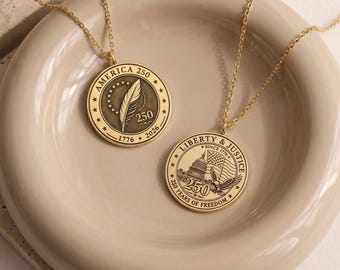 Op de afbeelding: Twee goudkleurige ronde hangers aan fijne kettinkjes. Eén hanger toont een veer, de tekst AMERICA 250 en de jaren 1776-2026. De andere hanger toont het Amerikaanse Capitool, een adelaar, een vlag en de tekst LIBERTY & JUSTICE.