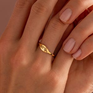 May include: A gold-colored ring with an oval face and delicate script. The ring is worn on a finger, showcasing its polished finish and intricate design. The ring's band is thin, and the script appears to be a word or phrase.