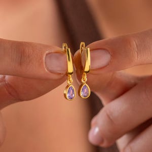 Op de afbeelding: Goudkleurige oorringen met druppelvormige paarse edelstenen. De oorbellen worden omhoog gehouden, waarbij het ontwerp en het contrast tussen het goud en de edelstenen worden getoond. De oorbellen zijn een sieraad.
