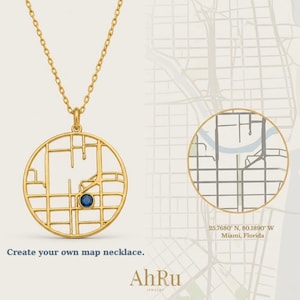 May include: A gold-colored necklace with a pendant shaped like a city map, featuring a blue gemstone. The necklace hangs on a delicate chain. The image includes the text "Create your own map necklace." and the brand name "AhRu Jewelry."