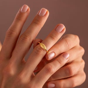 Può includere: Un anello dorato con il nome "Mary" inciso su una placca ovale. L'anello è indossato su un dito, mettendo in risalto il suo design delicato. L'anello è un gioiello.