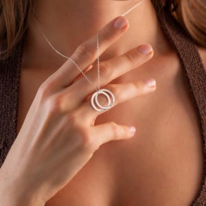 May include: A silver necklace featuring two interlocking circles. Each circle has a name engraved on it. The necklace is displayed on a person's hand, showcasing the delicate chain and pendant design. The background is a warm, neutral tone.