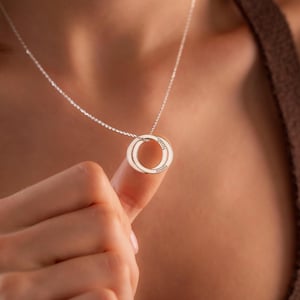 May include: A silver necklace with two intertwined rings. One ring is inscribed with the name "Noah", the other with "Amelia". The necklace is shown against a blurred background.