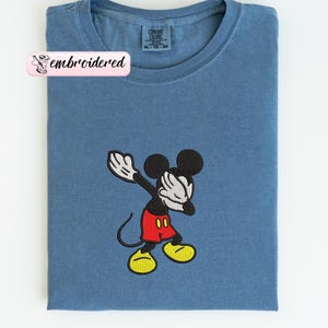 May include: A blue t-shirt with an embroidered Mickey Mouse design. The cartoon character is in a dabbing pose, with black ears, a black body, red shorts, yellow shoes, and white gloves. The shirt has a crew neck and a tag that says "Comfort Colors".