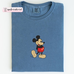 May include: A blue t-shirt with an embroidered image of Mickey Mouse. The cartoon character is wearing red shorts, yellow shoes, and a black jacket. The shirt has the text "embroidered" on a pink label.