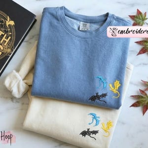 May include: Two sweatshirts, one light blue and one cream, are stacked. Each has three embroidered dragons in black, blue, and yellow. The light blue sweatshirt has the word "embroidered" in a pink banner. A book with gold lettering is visible.