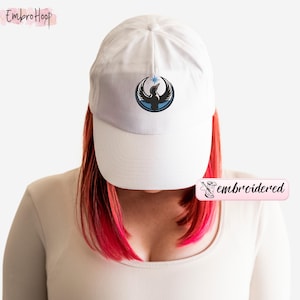 May include: White baseball cap with a curved brim. The cap features an embroidered design of a bird with a star in a blue circle. The word "embroidered" is printed on a pink label.