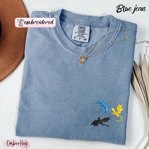 Pode incluir: Camiseta azul jeans com drag&otilde;es bordados em azul, amarelo e preto. A camisa tem gola redonda e mangas curtas. Um colar de ouro com um pingente est&aacute; drapeado sobre a camisa. O texto "Blue jean" est&aacute; no canto superior direito.