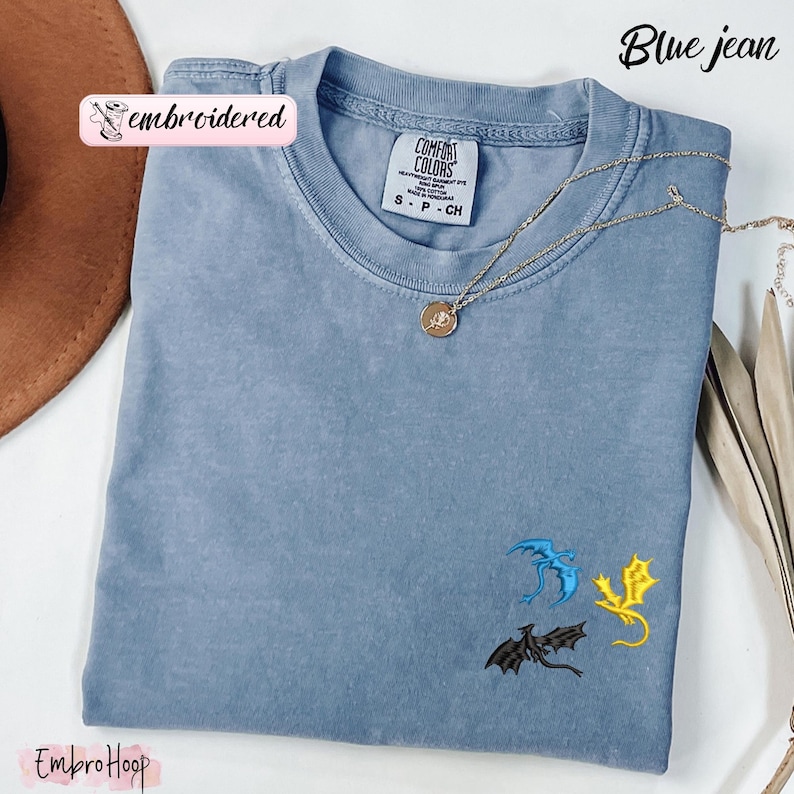 Pode incluir: Camiseta azul jeans com drag&otilde;es bordados em azul, amarelo e preto. A camisa tem gola redonda e mangas curtas. Um colar de ouro com um pingente est&aacute; drapeado sobre a camisa. O texto "Blue jean" est&aacute; no canto superior direito.