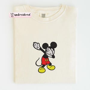 May include: Cream-colored t-shirt featuring an embroidered image of Mickey Mouse in a dabbing pose. Mickey is wearing red shorts, yellow shoes, and white gloves. The shirt has a Comfort Colors tag and a pink label that says "embroidered".