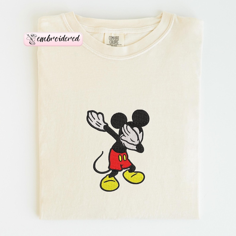 May include: Cream-colored t-shirt featuring an embroidered image of Mickey Mouse in a dabbing pose. Mickey is wearing red shorts, yellow shoes, and white gloves. The shirt has a Comfort Colors tag and a pink label that says "embroidered".