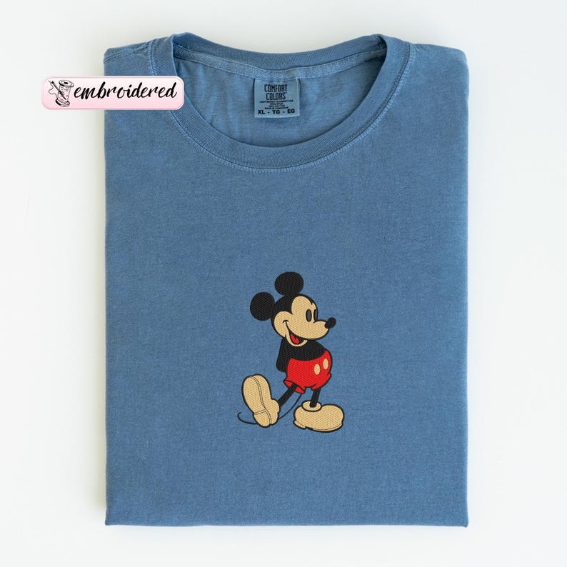 May include: A blue t-shirt with an embroidered image of Mickey Mouse. The cartoon character is wearing red shorts, yellow shoes, and a black jacket. The shirt has the text "embroidered" on a pink label.