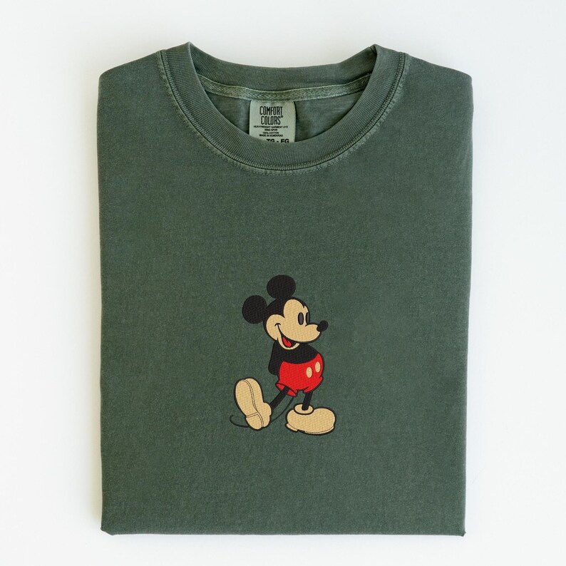 May include: Dark green t-shirt with a classic embroidered Mickey Mouse design. The shirt features a crew neck and short sleeves. The Mickey Mouse design includes black, red, and tan colors.
