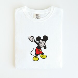 May include: White t-shirt featuring an embroidered image of Mickey Mouse dabbing. Mickey is depicted with black ears, arms, and face, red shorts, and yellow shoes. The shirt has a Comfort Colors tag.
