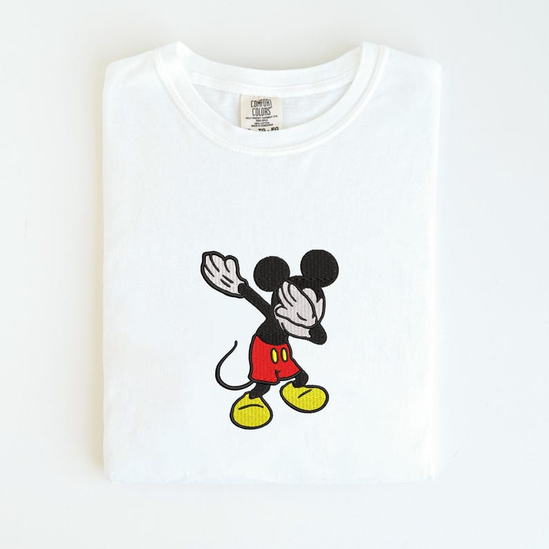 May include: White t-shirt featuring an embroidered image of Mickey Mouse dabbing. Mickey is depicted with black ears, arms, and face, red shorts, and yellow shoes. The shirt has a Comfort Colors tag.