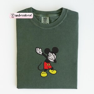May include: Dark green t-shirt featuring an embroidered image of Mickey Mouse in a dabbing pose. Mickey is wearing red shorts, yellow shoes, and white gloves. The shirt has a Comfort Colors tag and a pink label that says "embroidered".