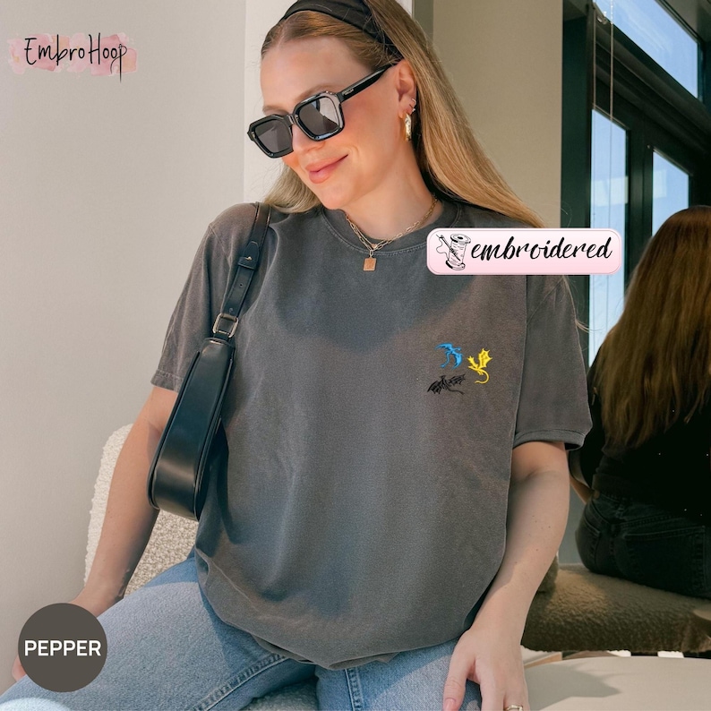 Pode incluir: Uma camiseta cinza com um pequeno desenho bordado de dois drag&otilde;es em azul e amarelo. A camiseta tem gola redonda e mangas curtas. A palavra "embroidered" est&aacute; impressa em uma etiqueta rosa.