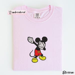 May include: A light pink Comfort Colors sweatshirt with an embroidered image of Mickey Mouse. Mickey is in a dabbing pose, wearing red shorts, yellow shoes, and white gloves. The word "embroidered" is in a pink banner.