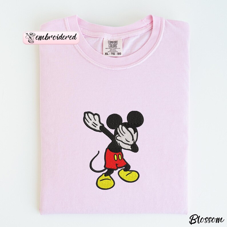 May include: A light pink Comfort Colors sweatshirt with an embroidered image of Mickey Mouse. Mickey is in a dabbing pose, wearing red shorts, yellow shoes, and white gloves. The word "embroidered" is in a pink banner.