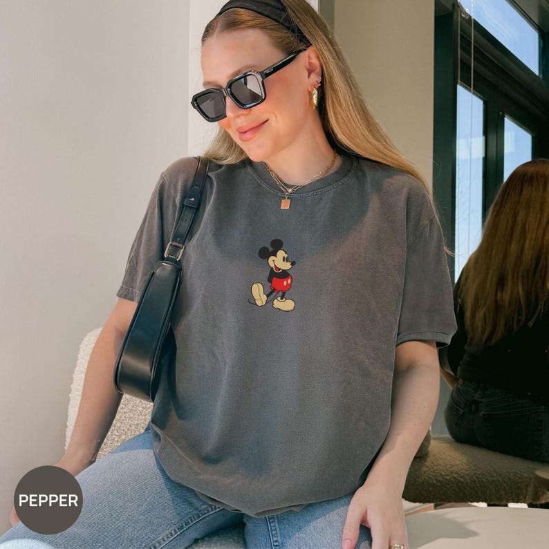 May include: A gray t-shirt featuring a classic Mickey Mouse graphic. The short-sleeved shirt has a crew neck and a slightly faded appearance. The graphic shows Mickey Mouse in his iconic pose, with red shorts and yellow shoes.