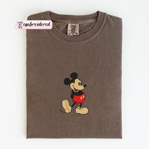 May include: A brown t-shirt featuring an embroidered Mickey Mouse design. The classic cartoon character is depicted in his signature red shorts, black top, and yellow shoes. The shirt has a crew neck and a tag that reads "Comfort Colors". The word "embroidered" is on a pink label.