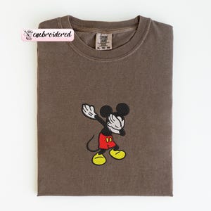 May include: A brown t-shirt with an embroidered Mickey Mouse design. Mickey is in a dabbing pose, wearing red shorts, yellow shoes, and white gloves. The shirt has a Comfort Colors tag and the word "embroidered" on a pink banner.