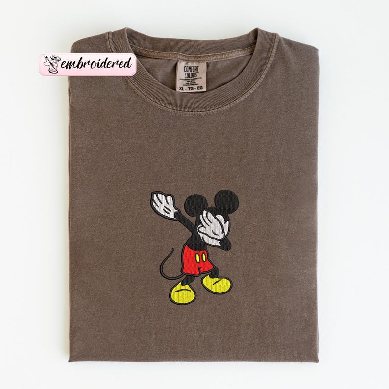 May include: A brown t-shirt with an embroidered Mickey Mouse design. Mickey is in a dabbing pose, wearing red shorts, yellow shoes, and white gloves. The shirt has a Comfort Colors tag and the word "embroidered" on a pink banner.