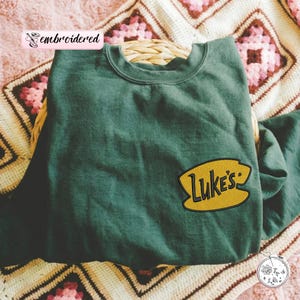 May include: A dark green sweatshirt with a yellow embroidered patch that reads "Luke's" in a stylized font. The sweatshirt is folded and resting on a woven basket, with a patterned blanket in the background.