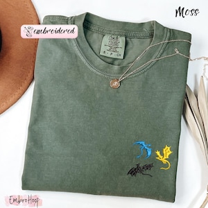 Pode incluir: Uma camiseta verde musgo com drag&otilde;es bordados em azul, amarelo e preto. A camiseta tem gola redonda e mangas curtas. Um colar de ouro com um pingente est&aacute; drapeado na camiseta. A palavra "embroidered" est&aacute; impressa em um adesivo rosa.