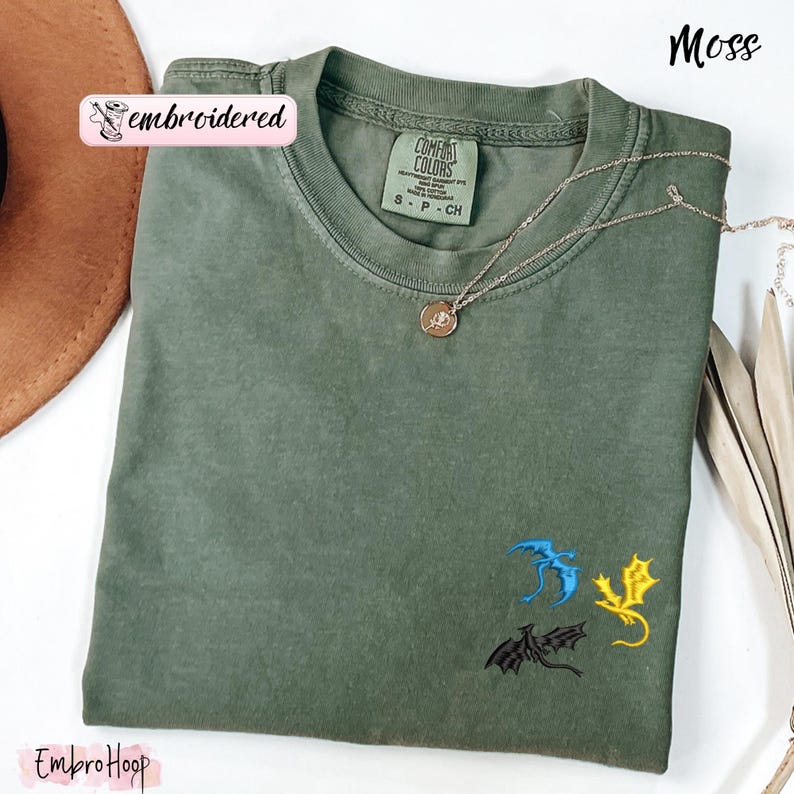 Pode incluir: Uma camiseta verde musgo com drag&otilde;es bordados em azul, amarelo e preto. A camiseta tem gola redonda e mangas curtas. Um colar de ouro com um pingente est&aacute; drapeado na camiseta. A palavra "embroidered" est&aacute; impressa em um adesivo rosa.
